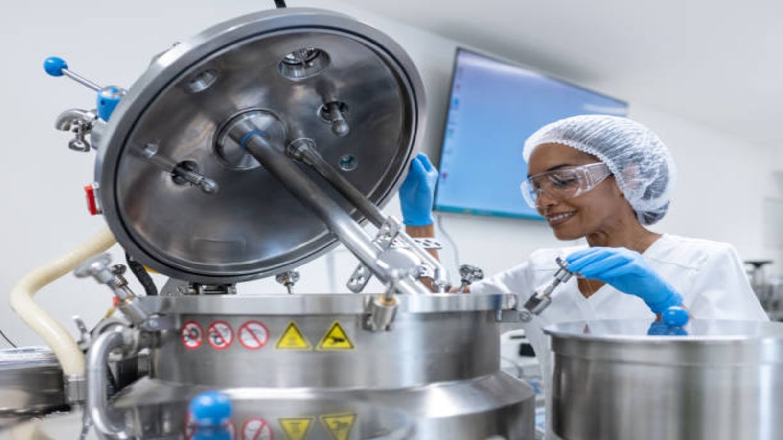 Multiracial group of female workers, pharmacists and researchers in biochemical pharmaceutical production during pharmaceutical manufacturing. A worker pours liquid into a homogenizer for the production of creams. The workers are in white protective suits, wear protective gloves, protective glasses and protective caps.